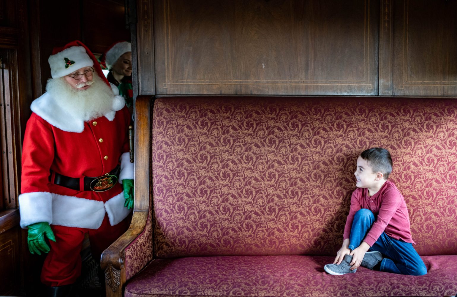 A small child sitting on a couch looking up at Santa Claus.